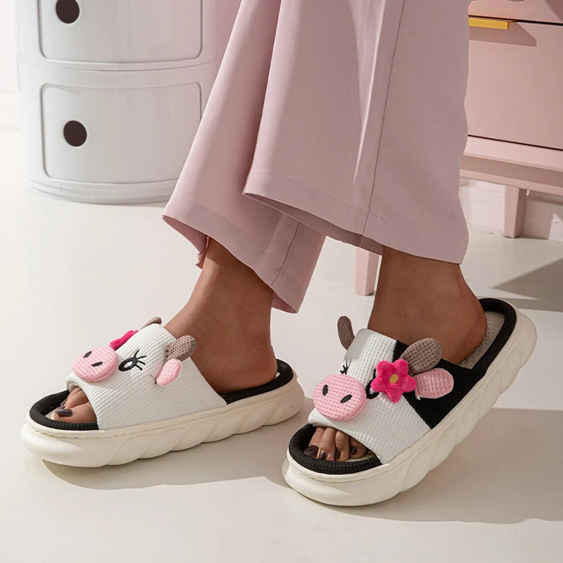 Pair of kawaii cartoon cow slippers with pink flowers worn by a person in light pink pants.