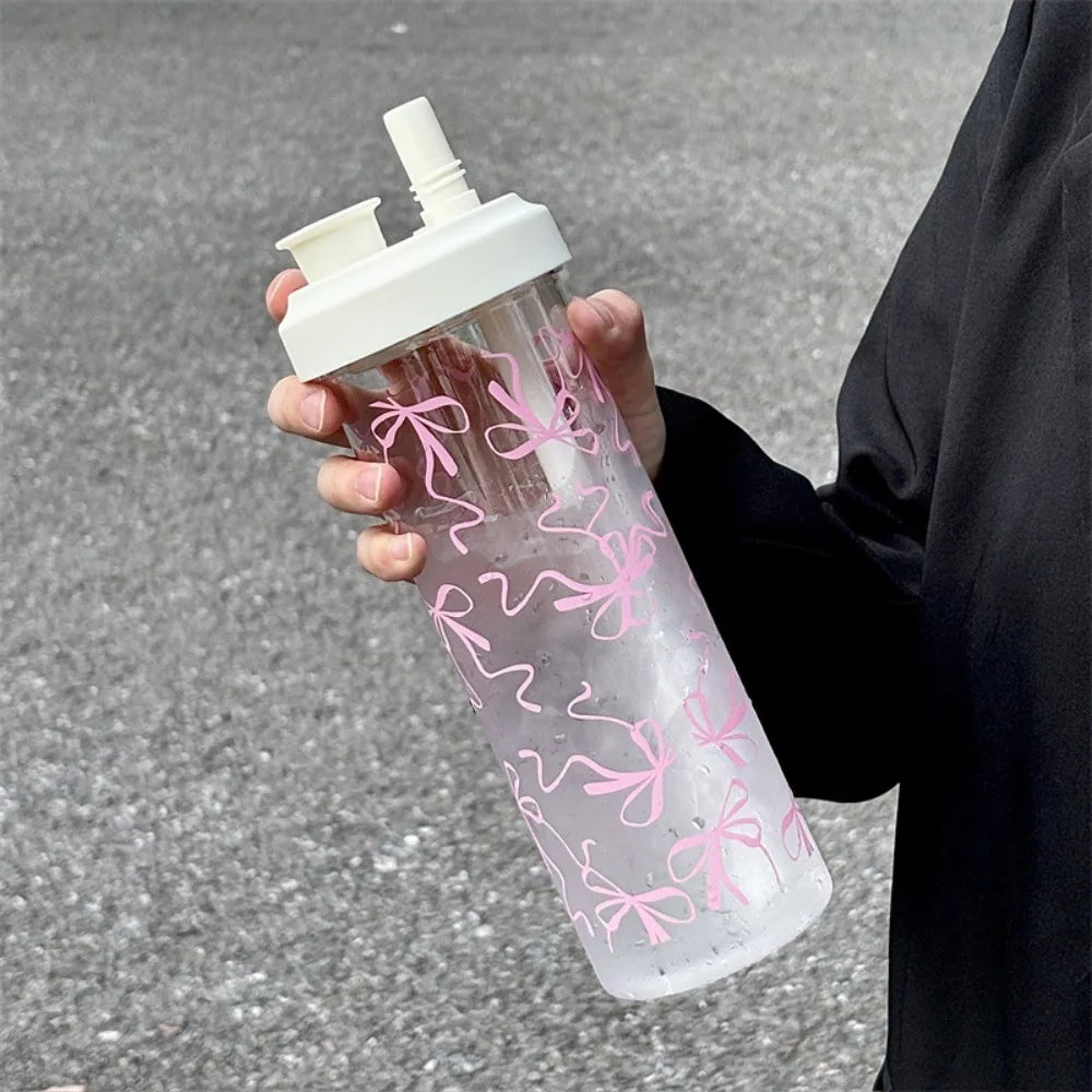 Clear Kawaii water bottle with pink bow designs held by a person on a gray pavement.