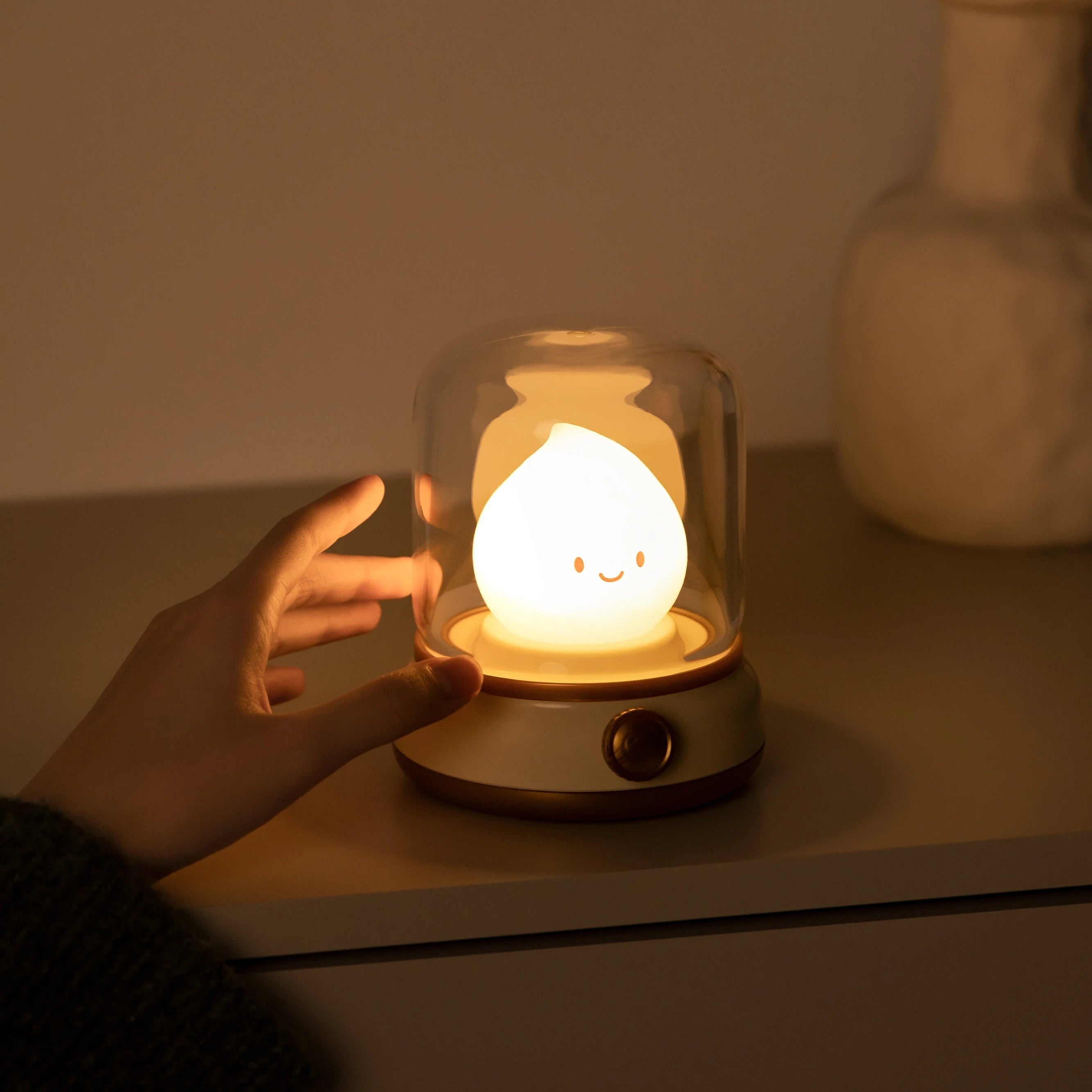 Hand holding a kawaii small lamp with a smiley face design under a glass dome on a wooden surface.