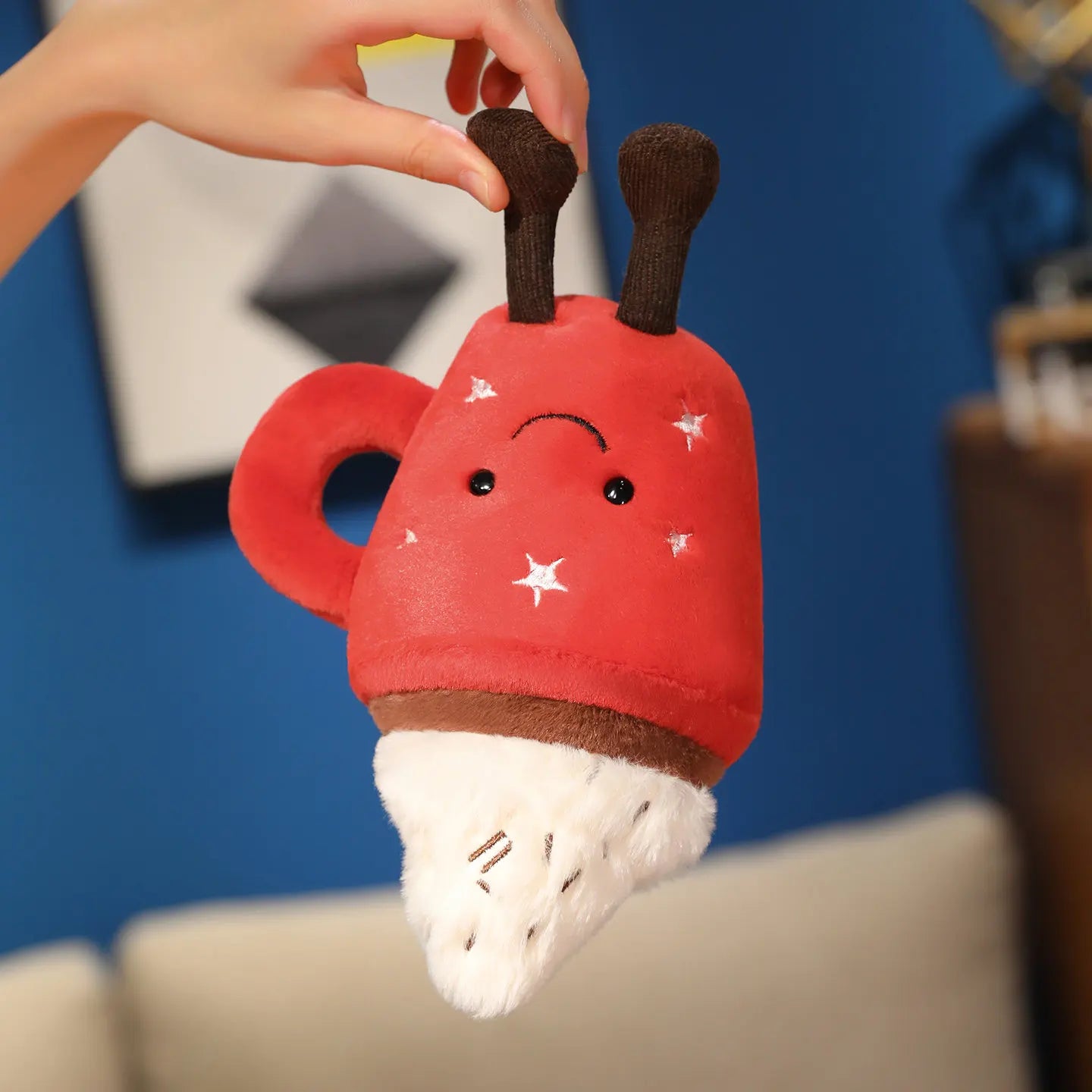 Red plush kawaii toy with brown antlers and white paws held by a hand against a blue wall.