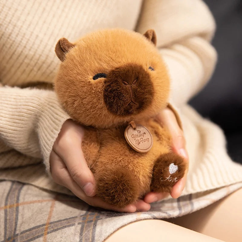 Person holding a small brown plush kawaii toy with a tag, sitting on a plaid blanket.
