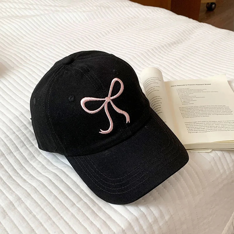 Black Kawaii cap with a pink bow on a white bed next to an open book