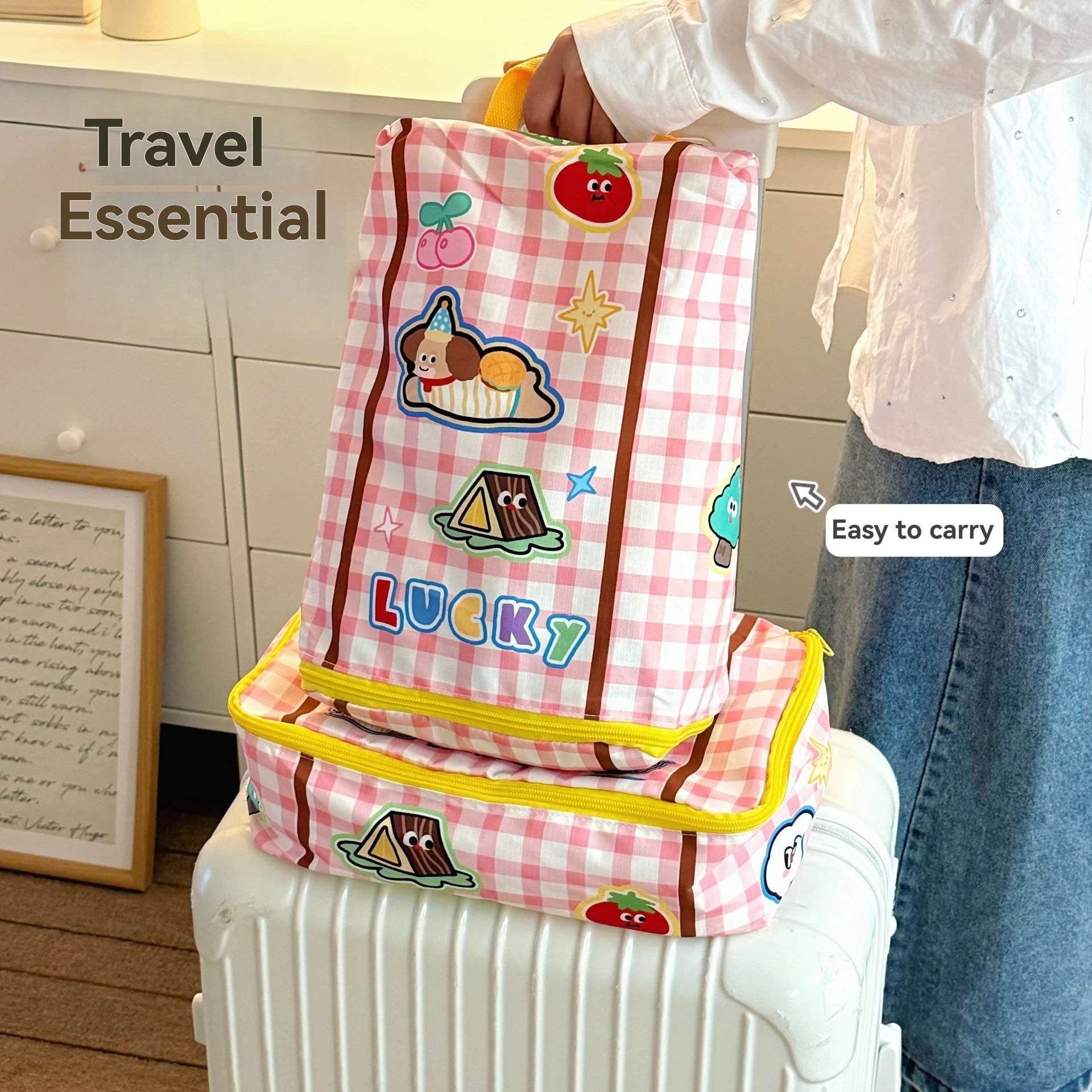 Pink checkered travel bag with Kawaii cartoon designs held by a person, placed on a white suitcase.