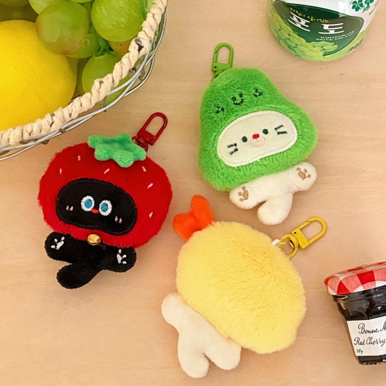 Three kawaii plush keychains shaped like a strawberry, avocado, and lemon on a wooden surface with fruits and a container in the background.