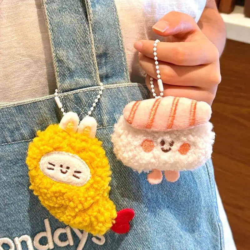 Two kawaii plush keychains, one yellow and one white with pink accents, held by a person wearing a denim apron.