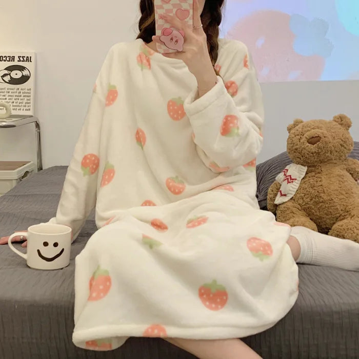 Person wearing a white fleece blanket with strawberry kawaii pattern, sitting on a bed with a teddy bear and a smiley face mug.
