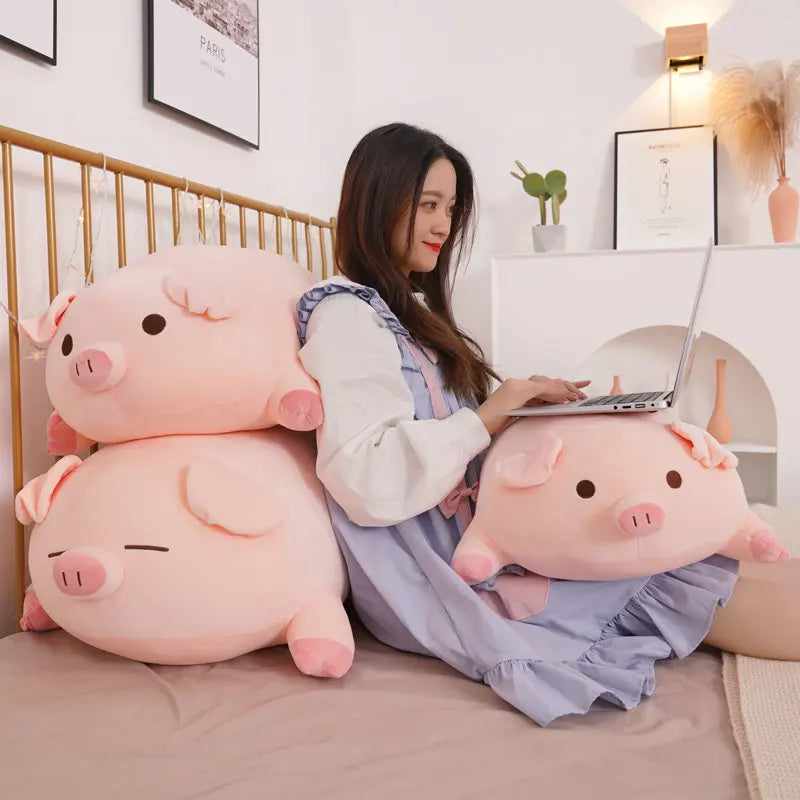 Woman sitting on a bed with two large pink pig-shaped kawaii cushions, using a laptop.