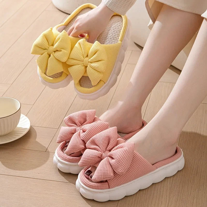 Yellow and pink slippers with large kawaii bows worn by a person on a wooden floor.