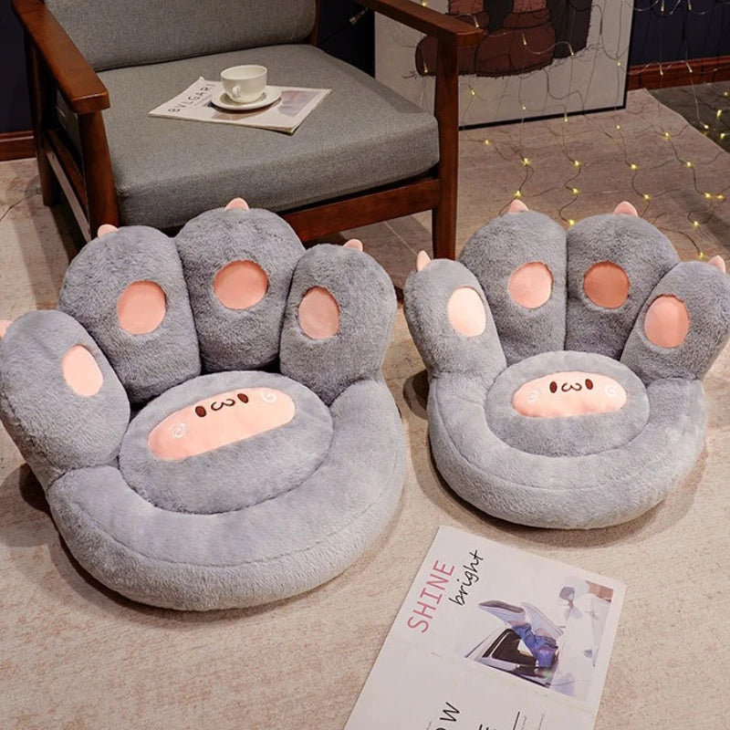 Two gray paw-shaped kawaii cushions on a floor with a chair and newspaper in the background.