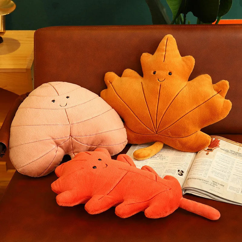 Three kawaii plush toys shaped like a pink mushroom, orange leaf, and red creature on a brown surface with an open book.