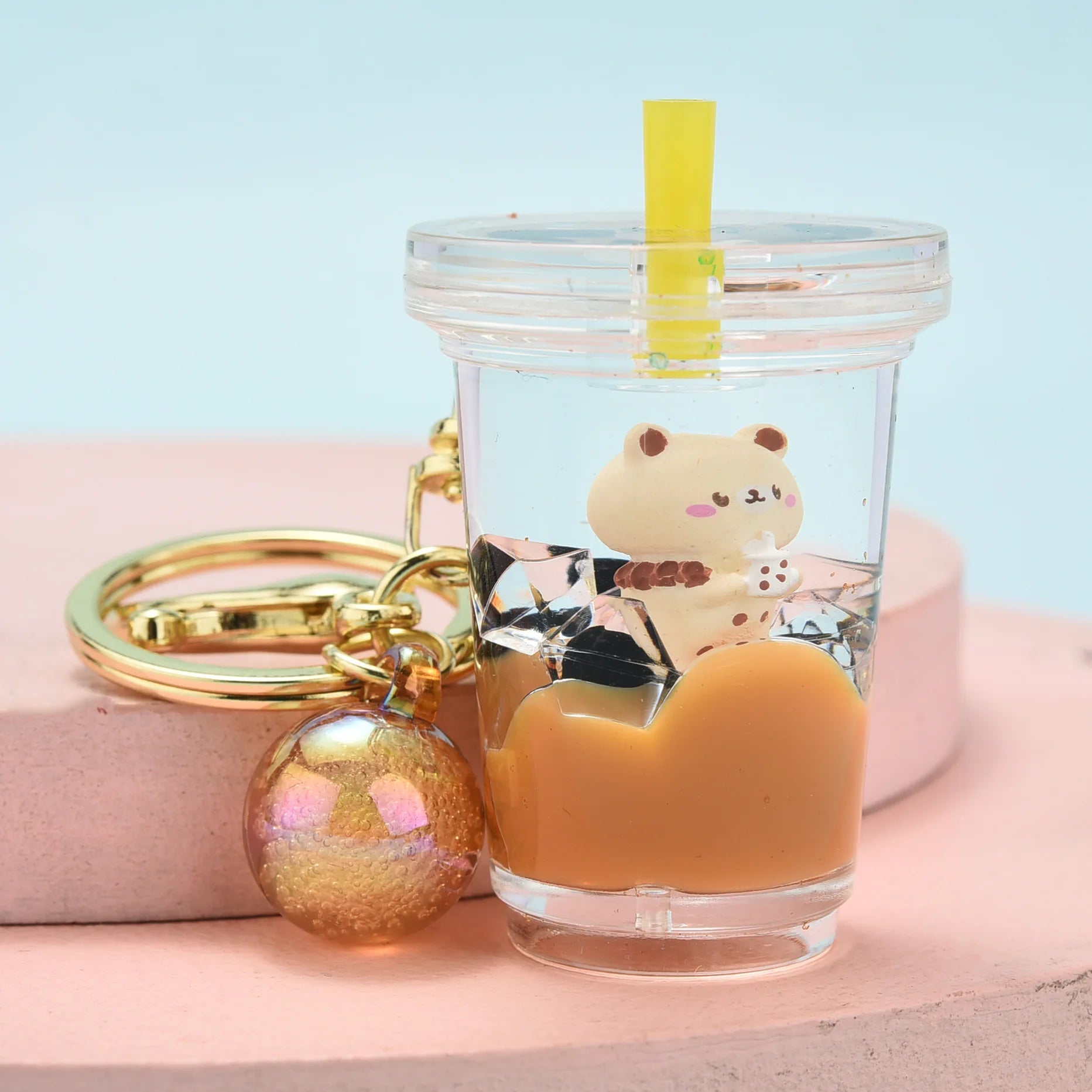 Clear tumbler with a kawaii bear charm, gold keychain, and orange fruit on a pink surface with a light blue background