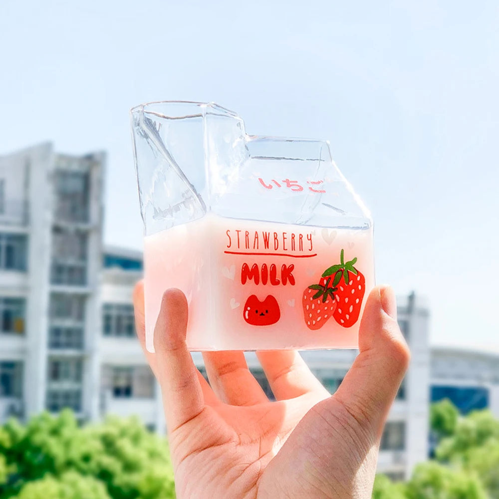 Hand holding a clear container with Kawaii strawberry milk design against a blurred outdoor background