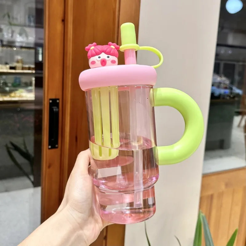 Pink and green Kawaii children's cup with handle and straw held by a hand indoors.