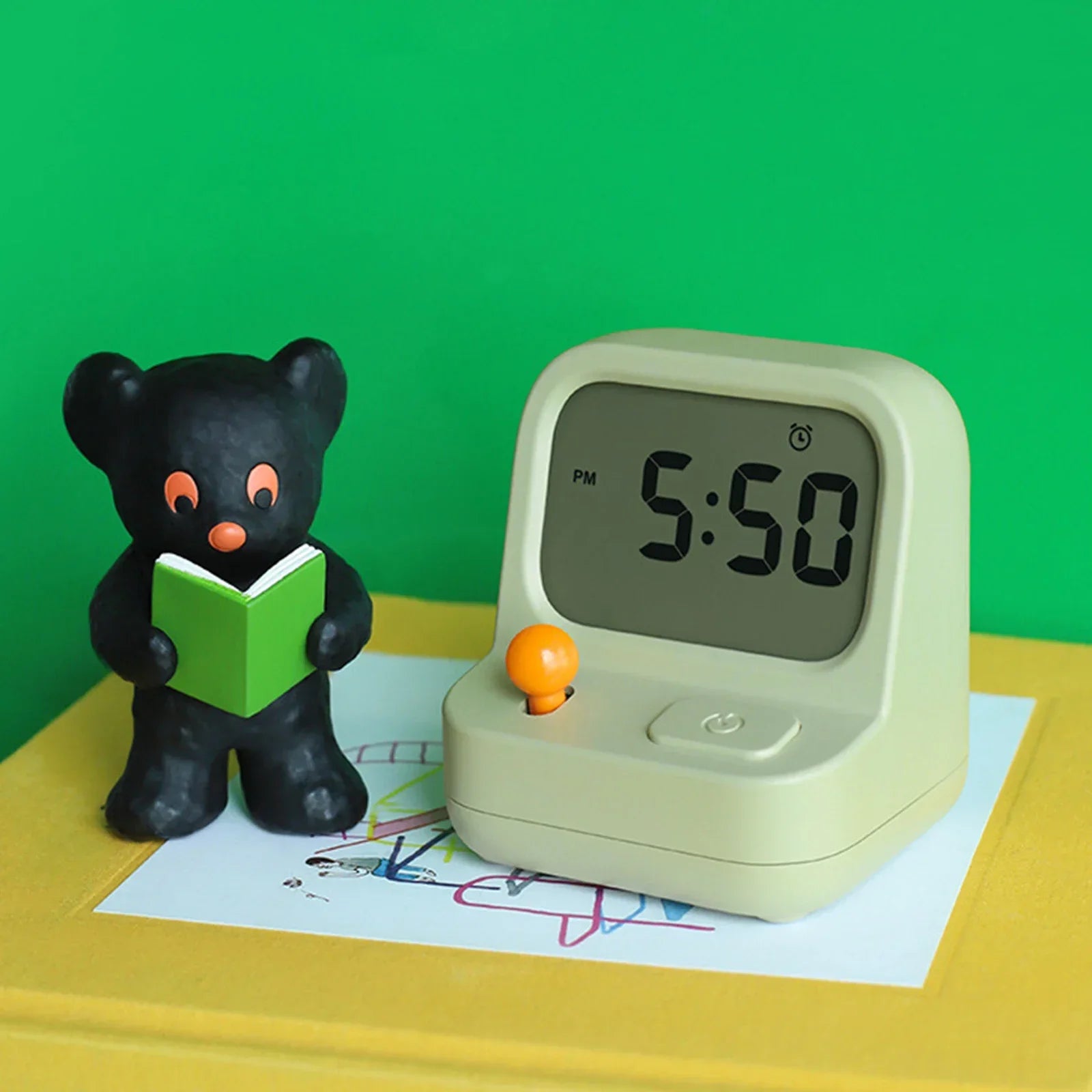 Small black bear figurine reading a book next to a kawaii digital clock on a yellow surface with a green background