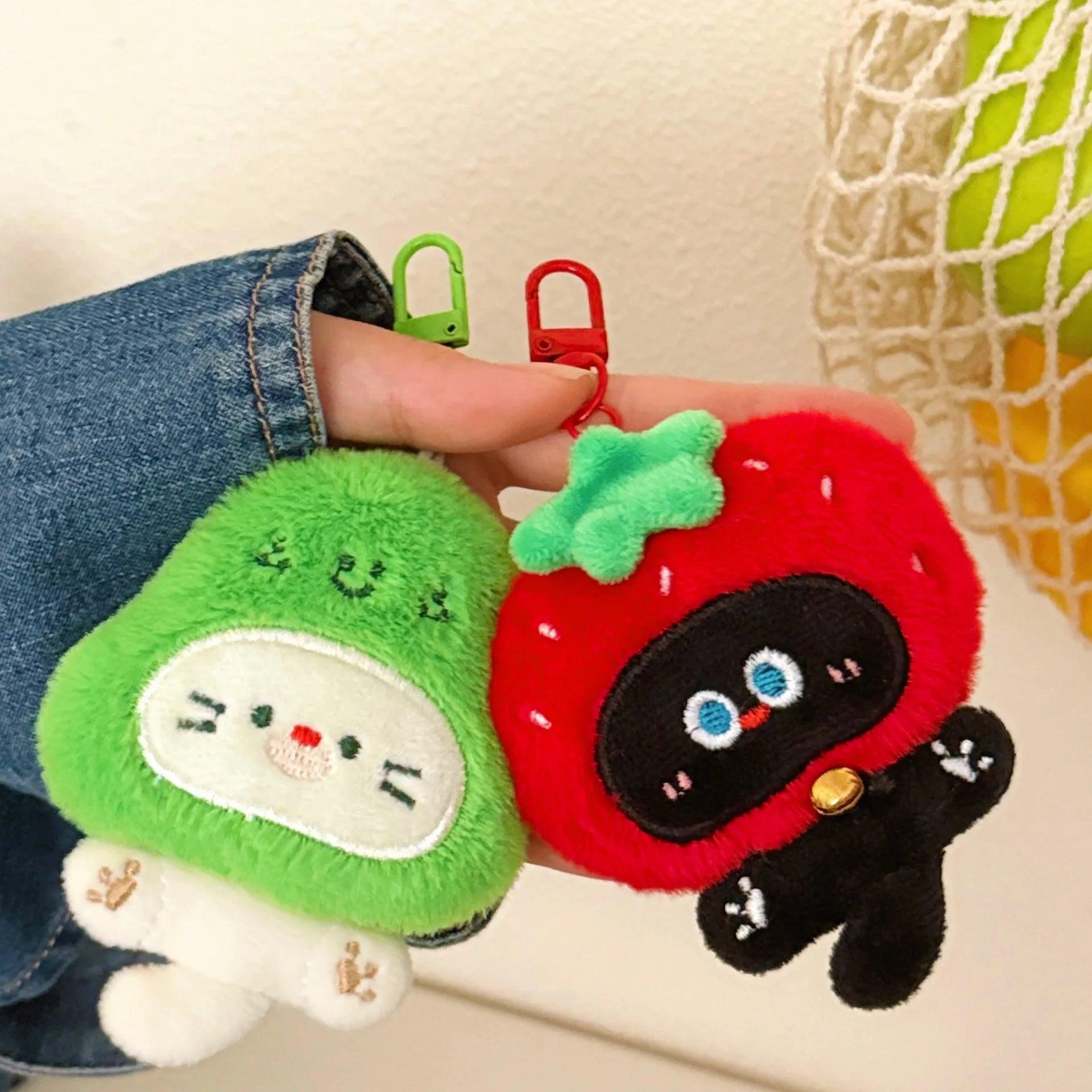 Two kawaii plush keychains, one green and one red with black accents, held by a person.