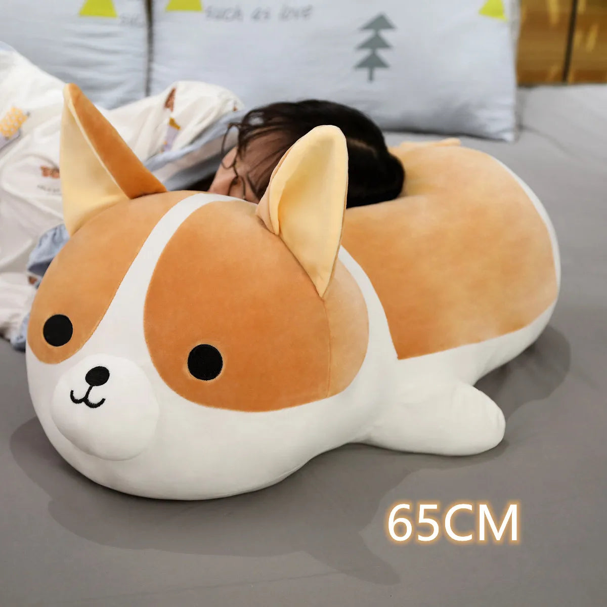 Corgi-shaped kawaii pillow on a bed with a person lying behind it, size indicated as 65CM.