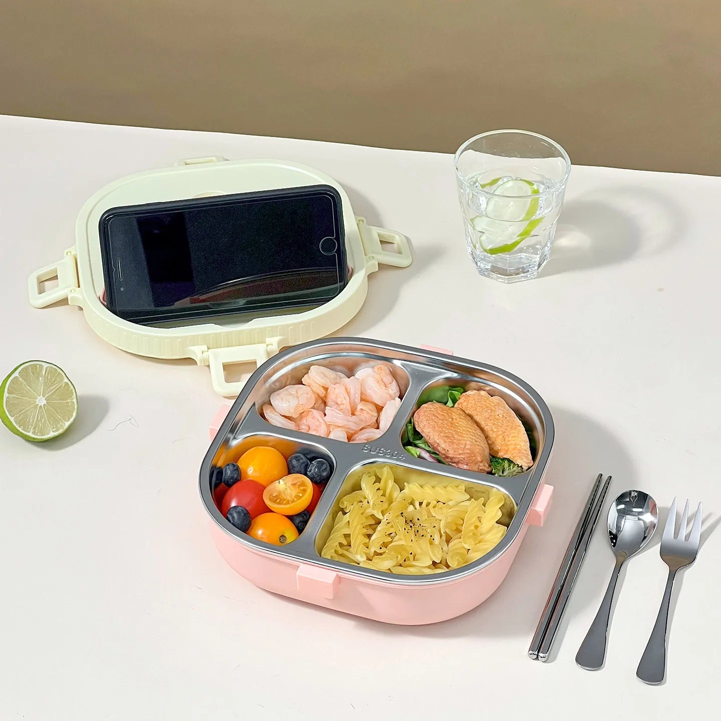 Pink kawaii bento box with compartments of food, smartphone, glass of water, and cutlery on a white surface.