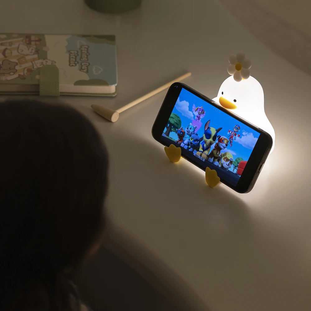 Kawaii phone holder shaped like a cartoon duck with a smartphone displaying a colorful app on a table.