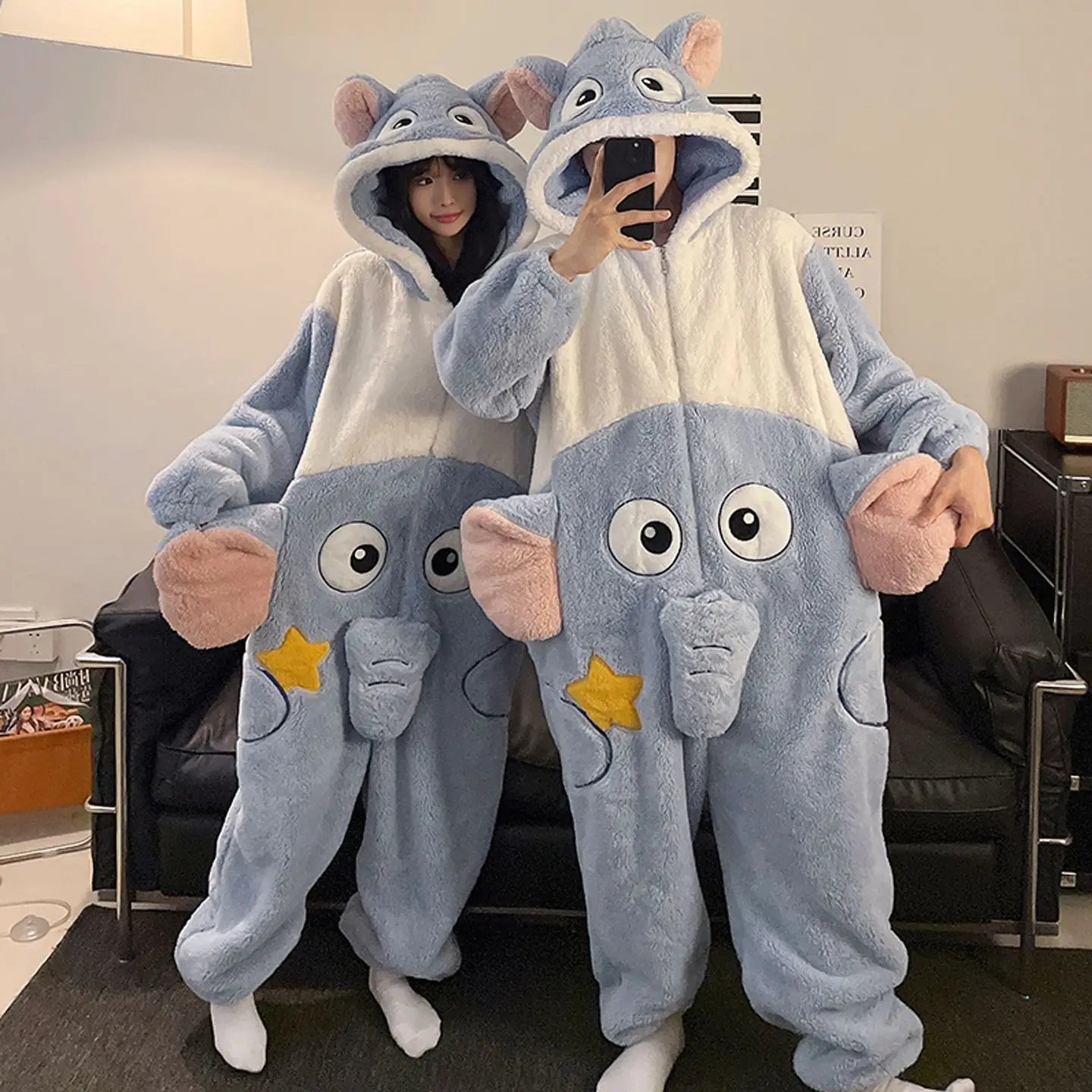 Two people wearing kawaii elephant-themed onesies taking a selfie in a living room.