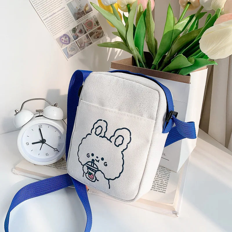 White bag with blue strap featuring a Kawaii cartoon design, placed on a surface with a clock and flowers in the background.