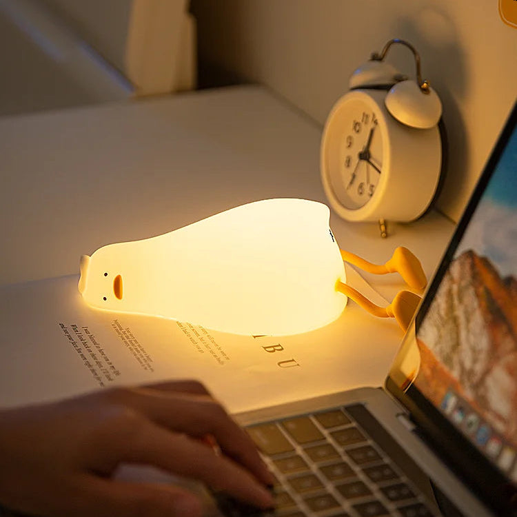 Person using a laptop with a glowing kawaii night light on an open book, next to a clock.