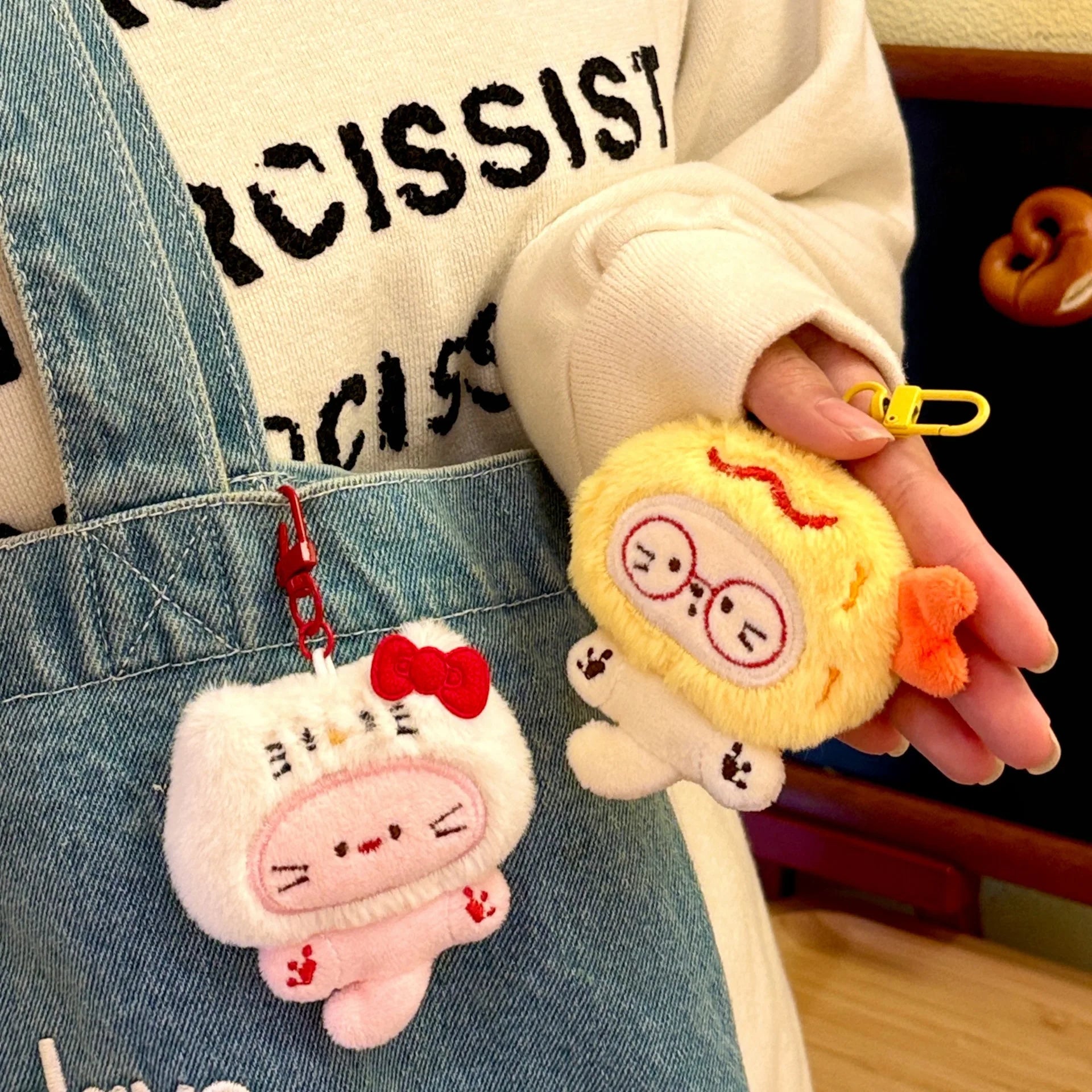 Two kawaii plush keychains attached to a denim overalls with a blurred background.