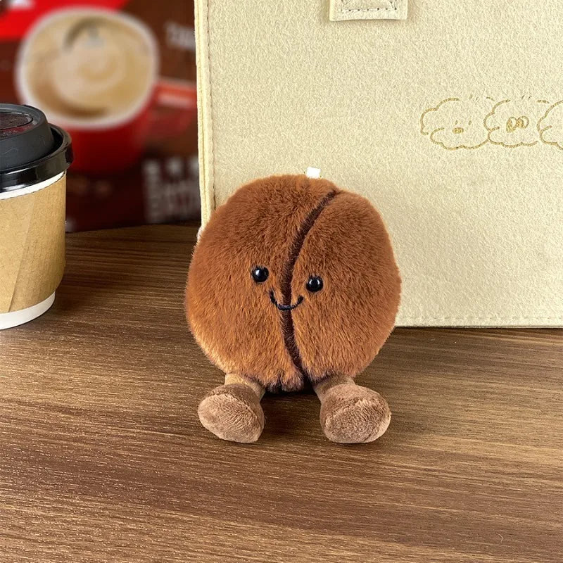 Plush kawaii toy shaped like a coffee bean on a wooden surface with a blurred coffee cup in the background.