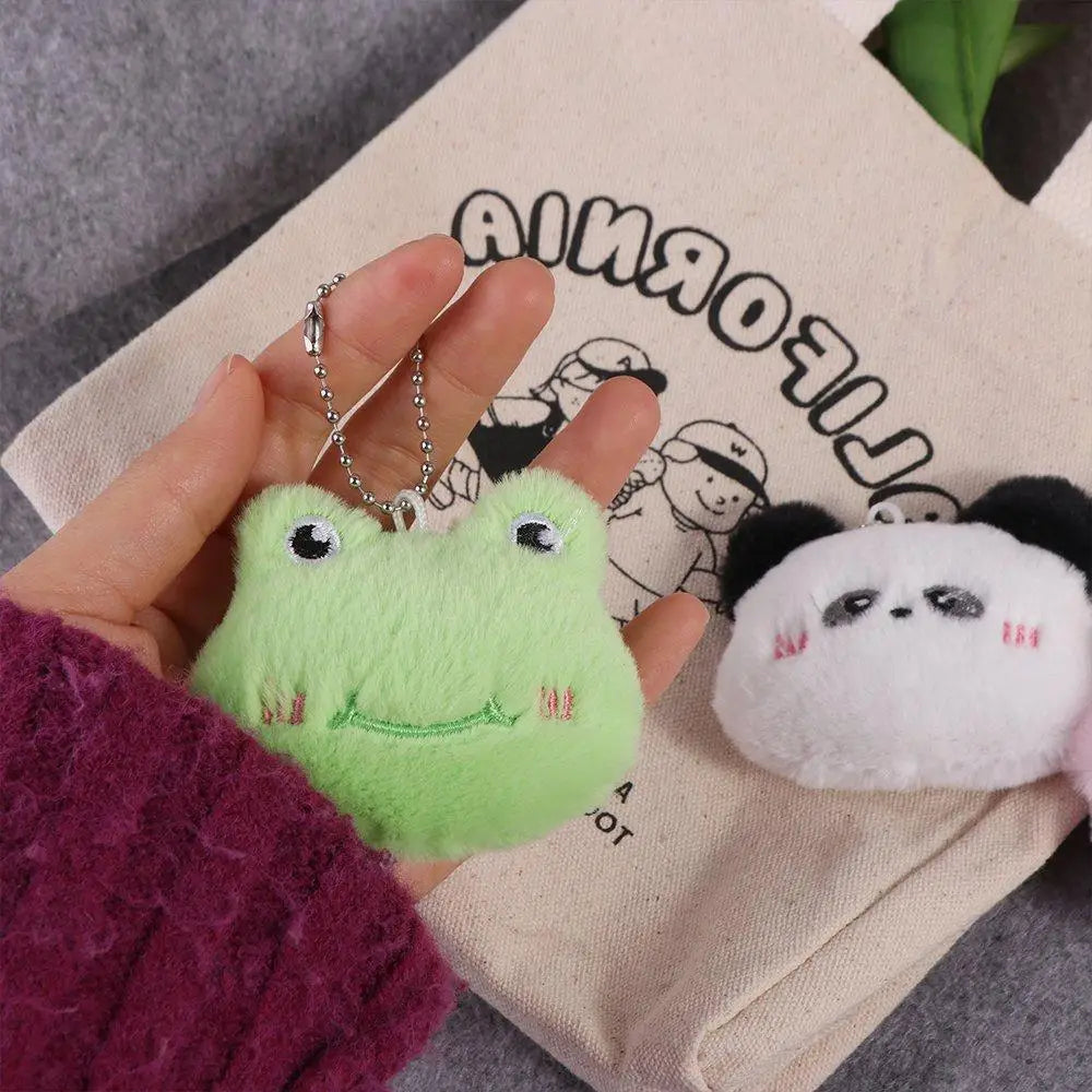 Hand holding a green kawaii plush frog keychain with a panda keychain in front of a beige bag with cartoon designs.