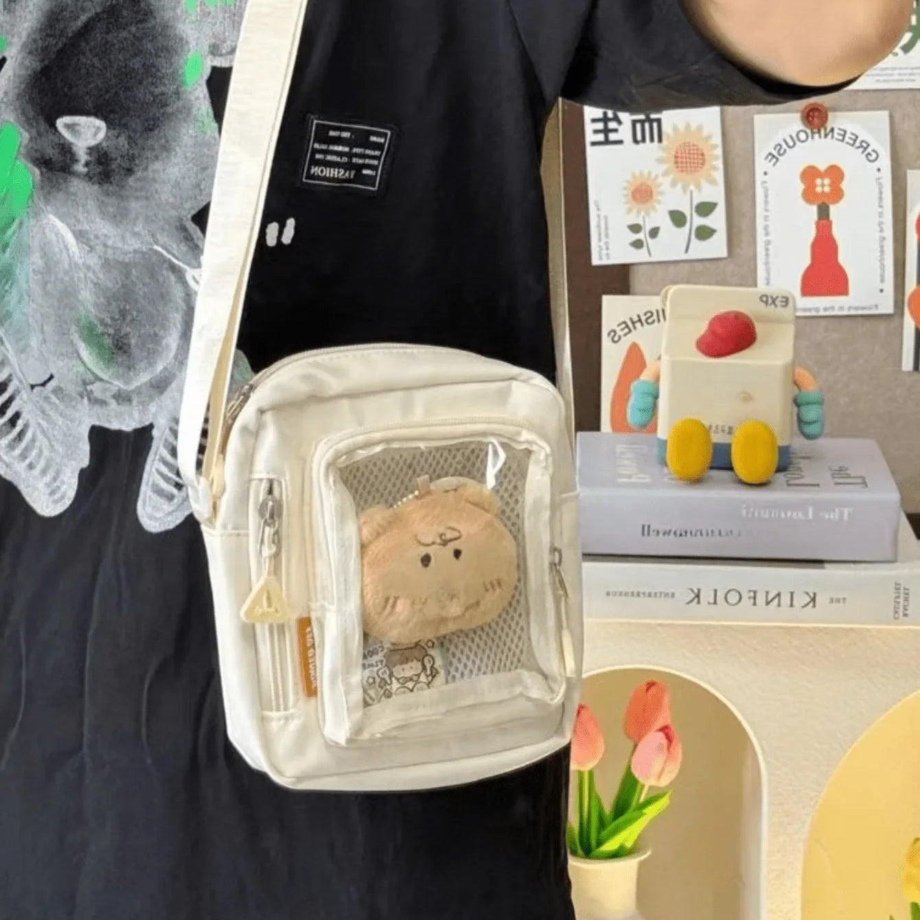 Person wearing a black shirt with a bear design, holding a small white bag with a Kawaii bear plush toy inside.