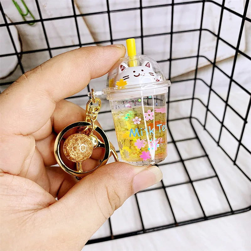 Hand holding a kawaii keychain shaped like a transparent cup with a cat design, filled with yellow liquid and colorful flowers.