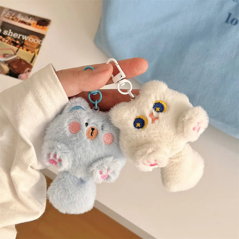 Two kawaii plush bear keychains, one gray and one white, held by a person with a magazine in the background.