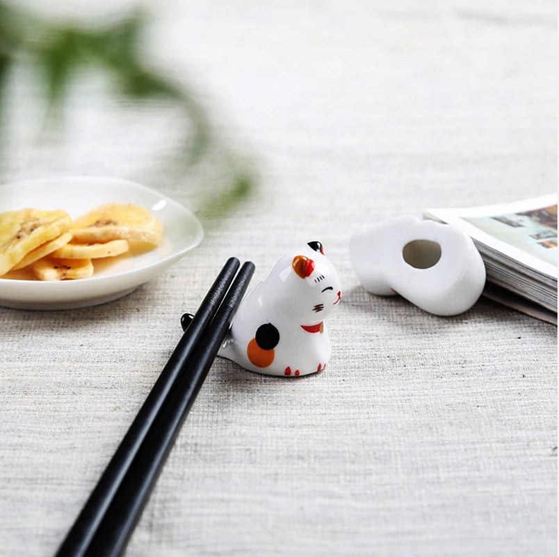White ceramic chopstick rest shaped like a kawaii cat with black and orange accents on a table with snacks and a magazine.