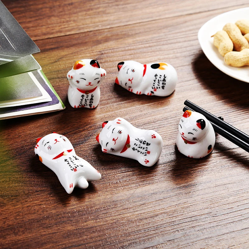 Five small ceramic kawaii cat figurines on a wooden table with a plate of snacks and chopsticks.