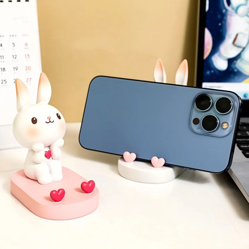 Kawaii phone stand shaped like a rabbit with pink hearts on a desk next to a smartphone.