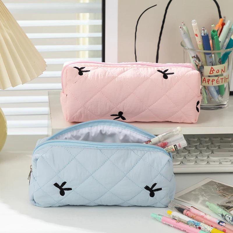 Two quilted kawaii pencil cases, one pink and one blue, on a desk with stationery items.