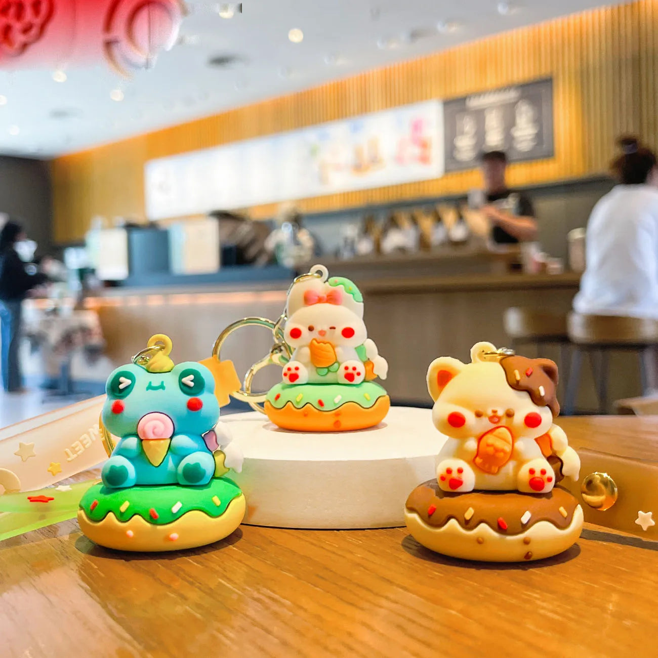 Three colorful kawaii animal-shaped keychains on a wooden table with a blurred restaurant interior in the background.
