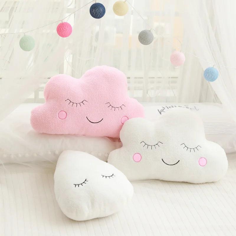 Set of cloud-shaped kawaii pillows with smiling faces on a soft surface with colorful polka dot string lights in the background.