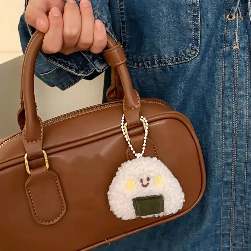 Brown handbag with a kawaii rice ball keychain attached, held by a person wearing blue jeans.
