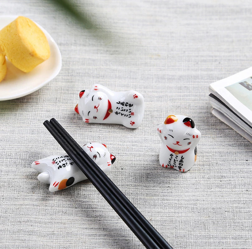 Three ceramic kawaii cat-shaped chopstick rests on a textured surface with a plate of fruit and book in the background.