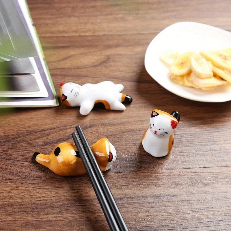 Three kawaii cat-shaped ceramic chopstick rest holders on a wooden surface with a plate of snacks in the background.